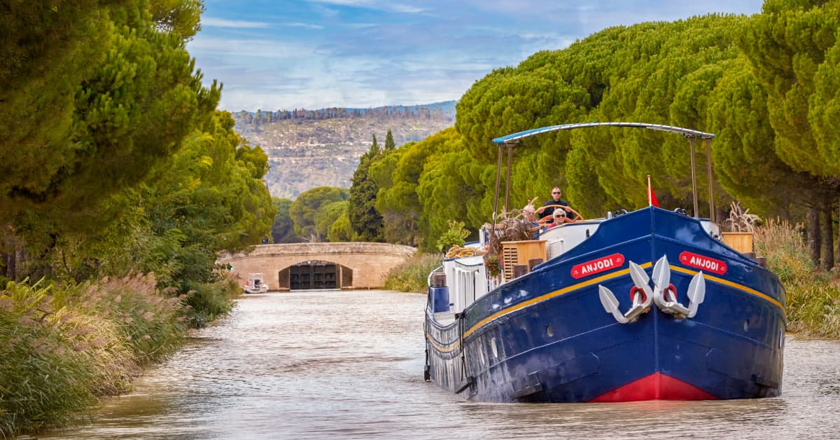 A traditional blue and gold barge named Anjodi navigates a picturesque tree-lined canal with