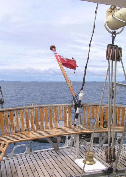 Sailing Vessel Deck with Red Flag at Sea A wooden deck of a sailing vessel with traditional rigging and pulleys visible, featuring a red