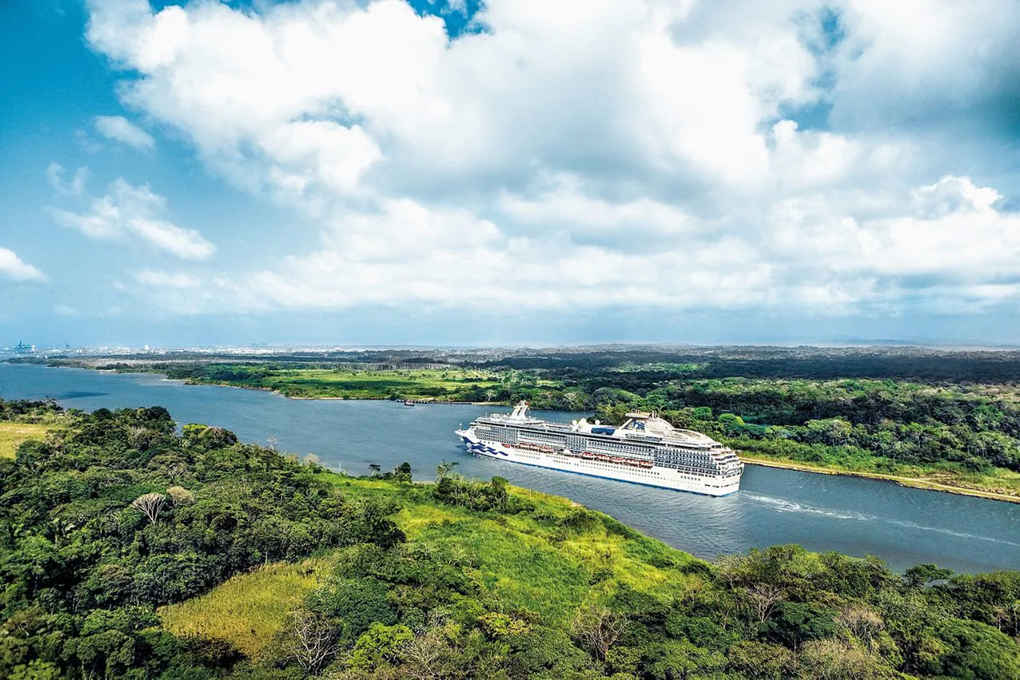 Cruise Ship Navigating Tropical Waterway A large modern cruise ship navigates through Gatun Lake surrounded by lush tropical rainforest and verdant vegetation. The image captures an aerial perspective of the vessel in the waterway with dense green foliage on both shorelines and a bright blue sky with white clouds overhead.