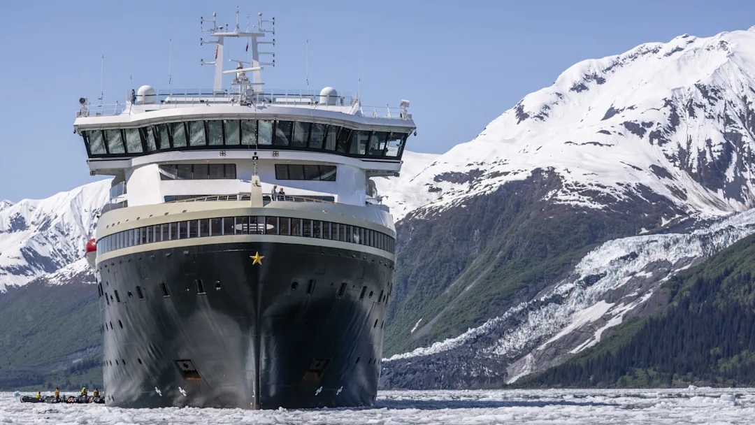 MS Roald Amundsen in College Fjord, Alaska A large cruise ship sailing through icy waters with snow-capped mountains and glacial terrain in the background. A small zodiac boat is visible near the ship's hull.