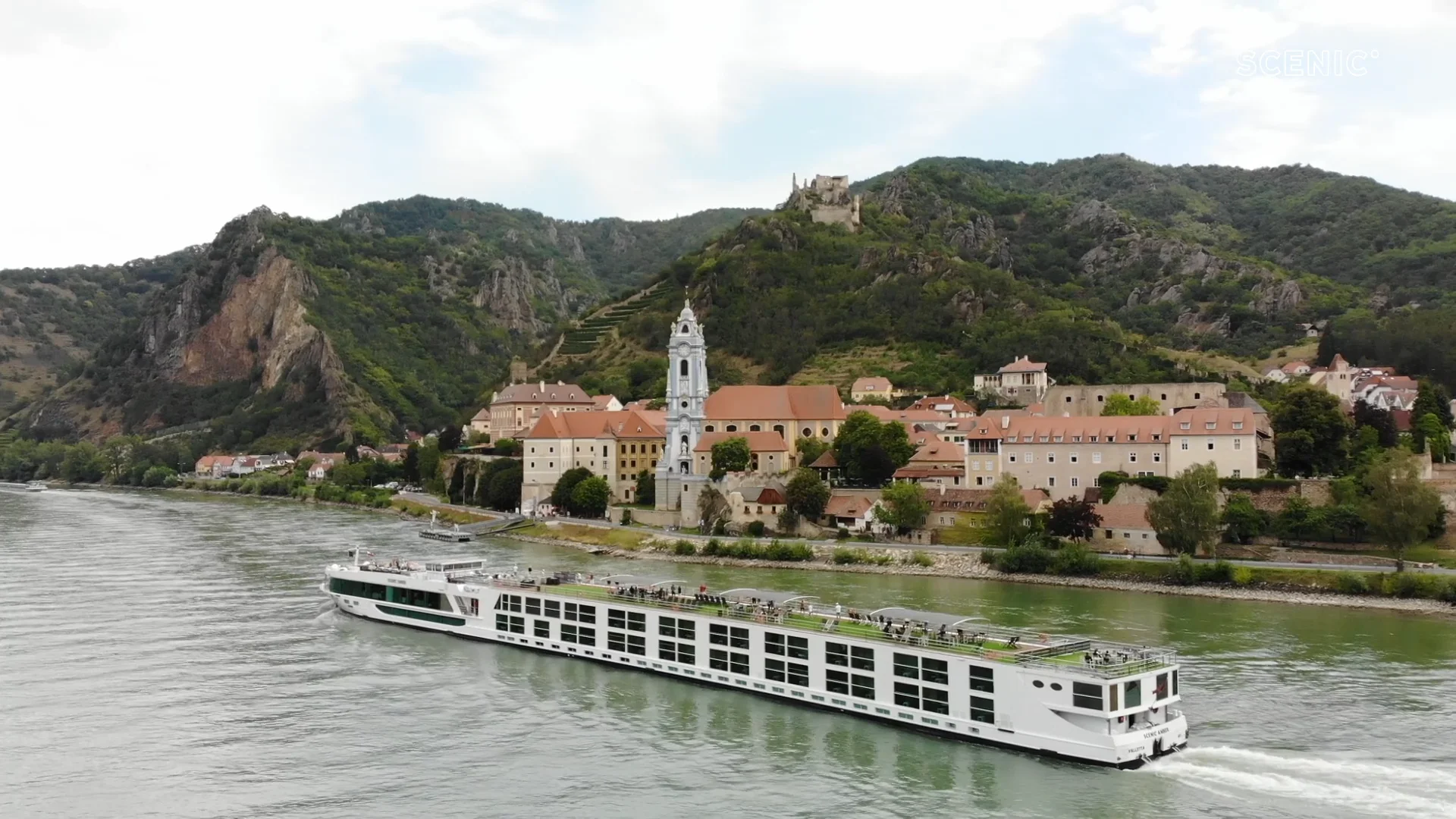 A modern river cruise ship navigates the Danube River past a picturesque medieval town nestled in