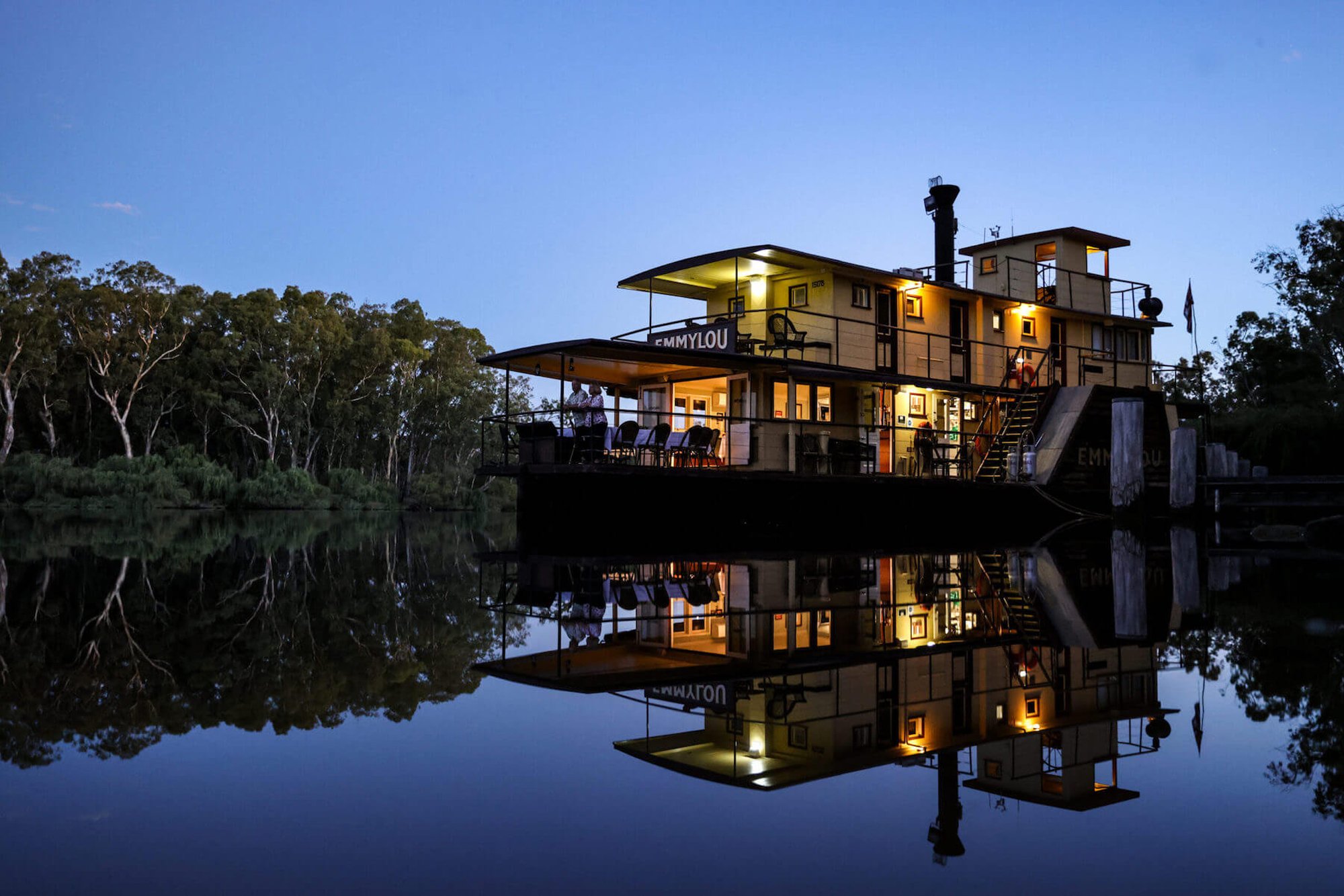 Emmylou Riverboat at Night with Reflections A beautifully illuminated multi-level houseboat named Emmylou glows warmly against the twilight sky, its reflection perfectly mirrored in the still, dark waters below. Surrounded by eucalyptus trees along a serene riverbank, the vessel showcases modern architecture with multiple decks, large windows, and outdoor living spaces.