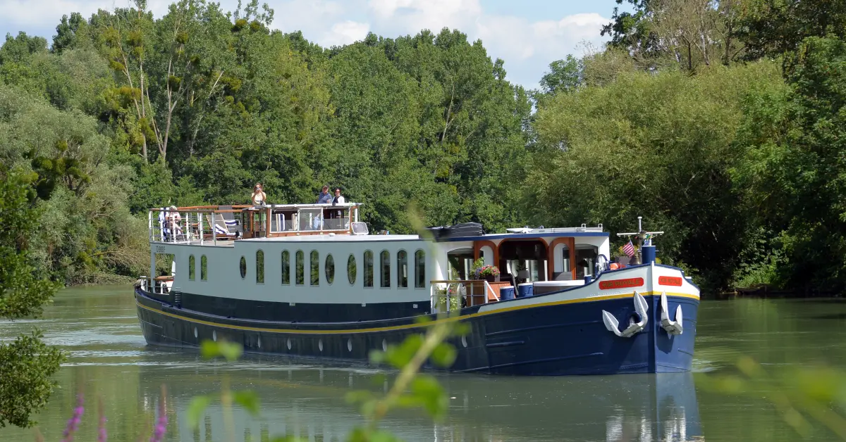 A elegant modern barge with white upper decks and dark blue hull navigates a serene river lined