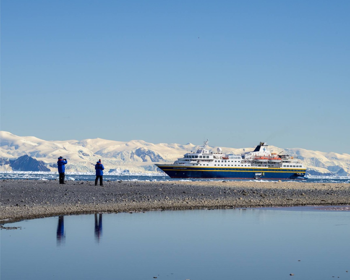 Expedition Cruise Ship at Arctic Beach A large expedition cruise ship with blue and white hull anchors offshore at a rocky Arctic beach