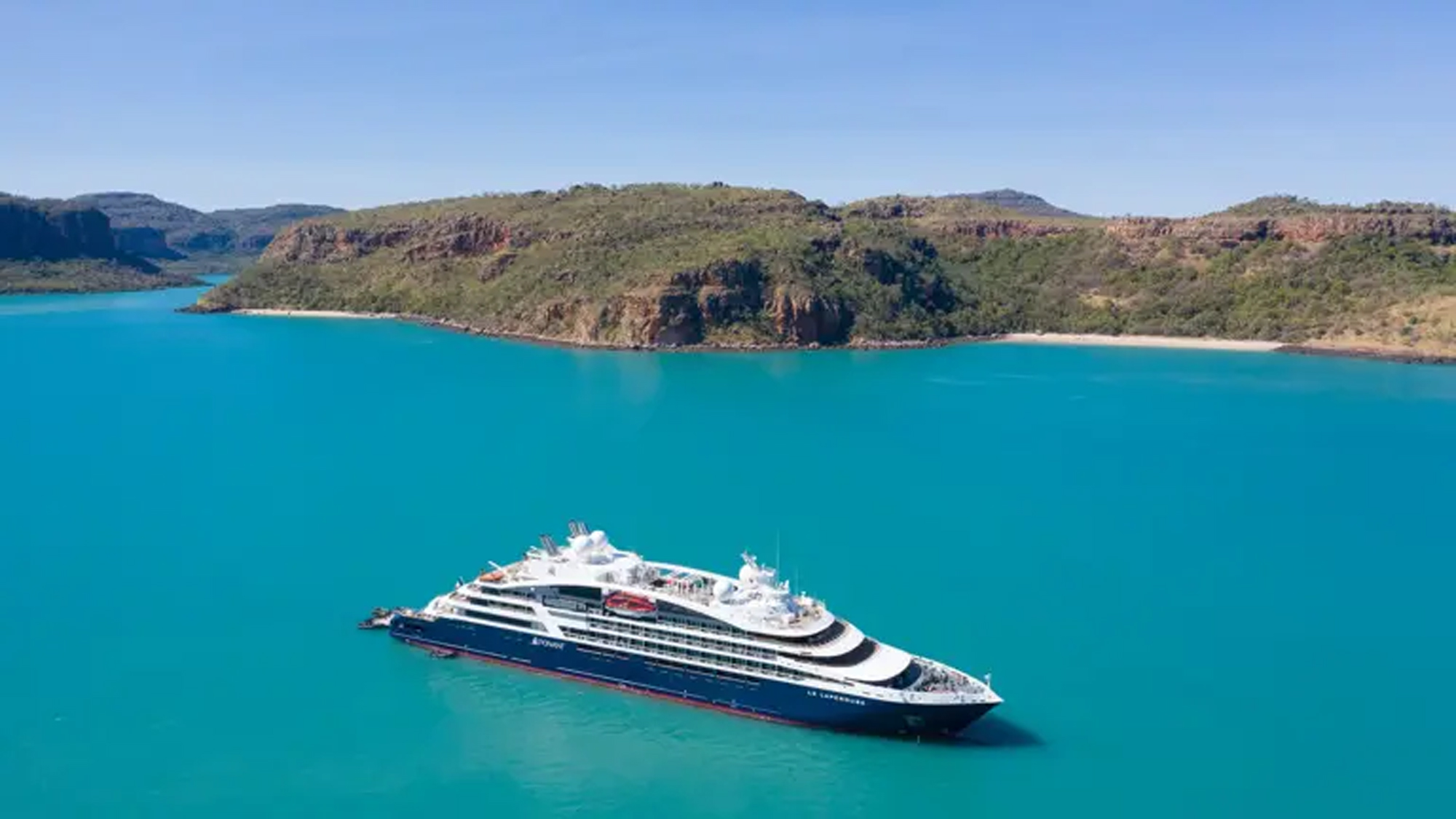 Luxury Cruise Ship in Turquoise Waters A modern expedition cruise ship anchors in pristine turquoise waters off a rugged, vegetation-covered peninsula in Australia's remote Kimberley region. The dramatic landscape features rocky outcrops, dense bushland, and a secluded white sandy beach, exemplifying the untamed natural beauty of this World Heritage area.