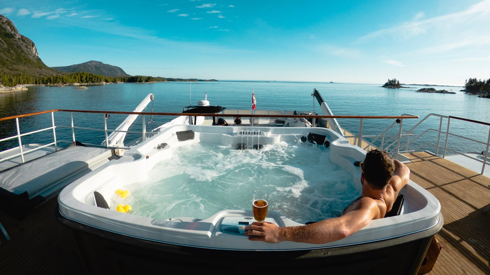 Luxury Hot Tub on Expedition Yacht Deck A person relaxes in a large, bubbling hot tub on the deck of a luxury yacht, holding a beverage while enjoying panoramic views of a pristine turquoise lake surrounded by forested mountains and rocky shores.