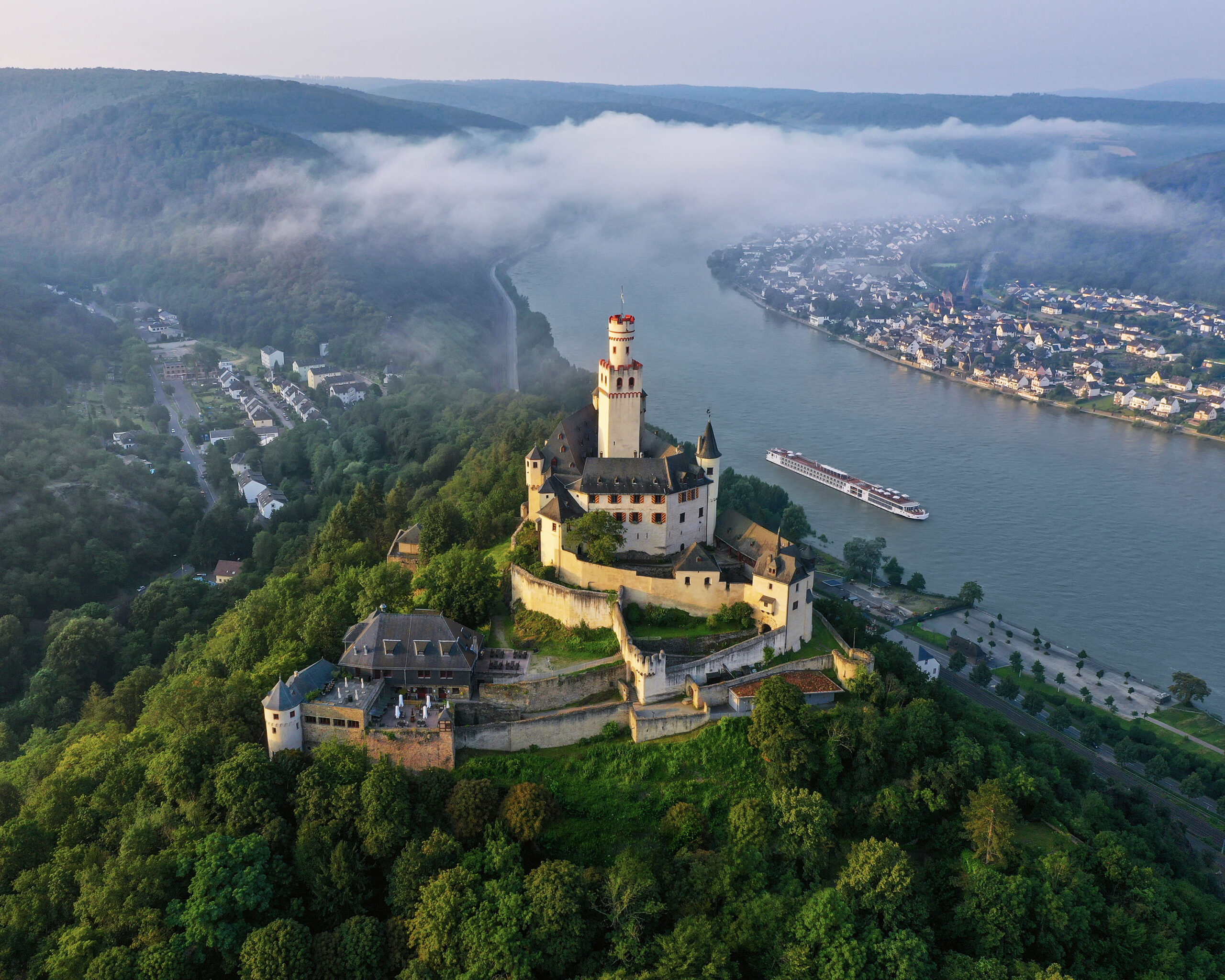 Marksburg Castle Above Rhine River Valley An aerial view of the iconic Neuschwanstein Castle perched on a hilltop above the Rhine River in Germany, with a river cruise ship navigating the misty valley below. The castle's distinctive yellow limestone walls and towers rise majestically from dense forests, while morning fog blankets the picturesque riverside towns and vineyards along the river bend.
