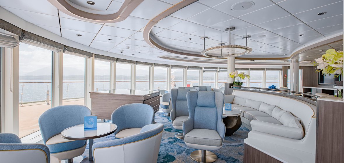 Modern Cruise Ship Lounge with Ocean Views A sophisticated and contemporary lounge area aboard a luxury cruise ship featuring floor-to-ceiling windows with panoramic ocean views, stylish blue and gray upholstered seating, modern pendant lighting, and elegant architectural details including curved ceiling elements and wooden accents throughout the space.