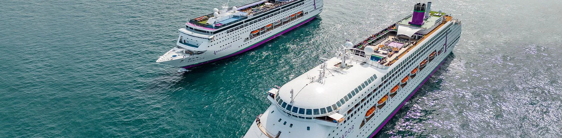 Multiple Cruise Ships Docked at Port A large modern cruise ship named Ambassador, featuring white hull with distinctive purple and orange accent bands, docked at a tropical port with clear blue waters. The vessel displays multiple decks, communication satellites, and lifeboats typical of contemporary ocean liner design.