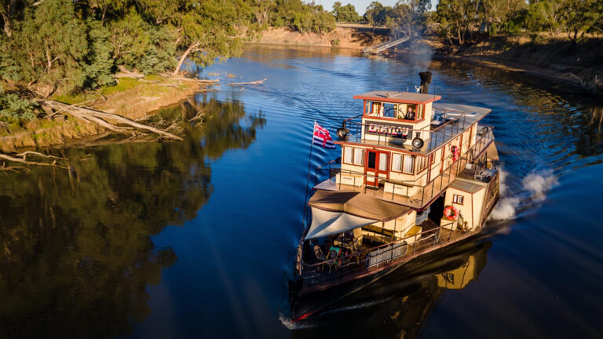 Paddlewheel Riverboat on Murray River A classic multi-deck paddlewheel riverboat with cream and red exterior navigates calm waters lined