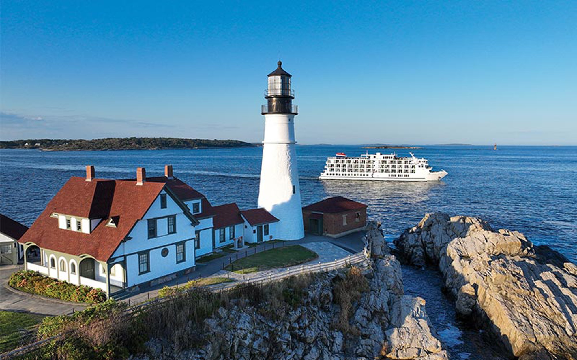 Portland Head Light with Cruise Ship A scenic coastal view of the iconic Portland Head Light lighthouse with its distinctive white tower and black top, accompanied by a charming blue keeper's house and supporting buildings perched on rocky cliffs. A modern cruise ship can be seen anchored in the calm blue waters offshore, showcasing this popular New England cruise destination.