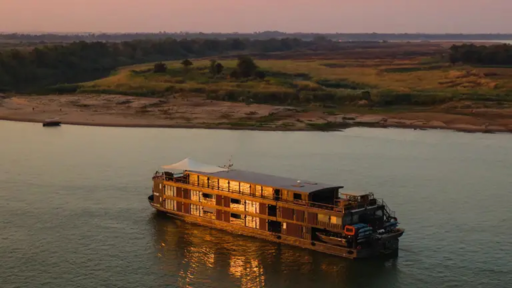 River Cruise Boat at Golden Hour A traditional wooden river cruise ship navigates the Mekong River at sunset, with its distinctive multi-deck design illuminated by golden light. The vessel is positioned against a scenic backdrop of rural floodplain landscape with scattered trees and distant forests under a warm, hazy sky.