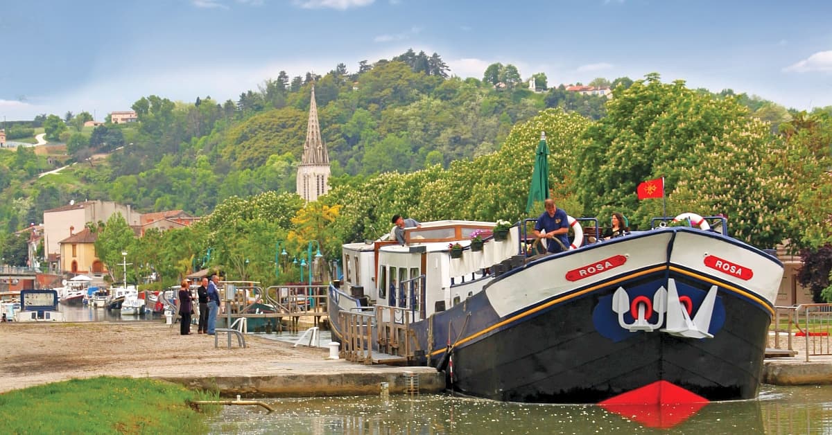 ROSA River Cruise Ship Docked in European Town The river cruise ship Rosa is moored at a picturesque European riverport with passengers on deck, surrounded by lush green hillsides, a church spire visible in the background, and traditional riverside architecture. The scene captures the essence of leisurely river cruising through a charming continental landscape.