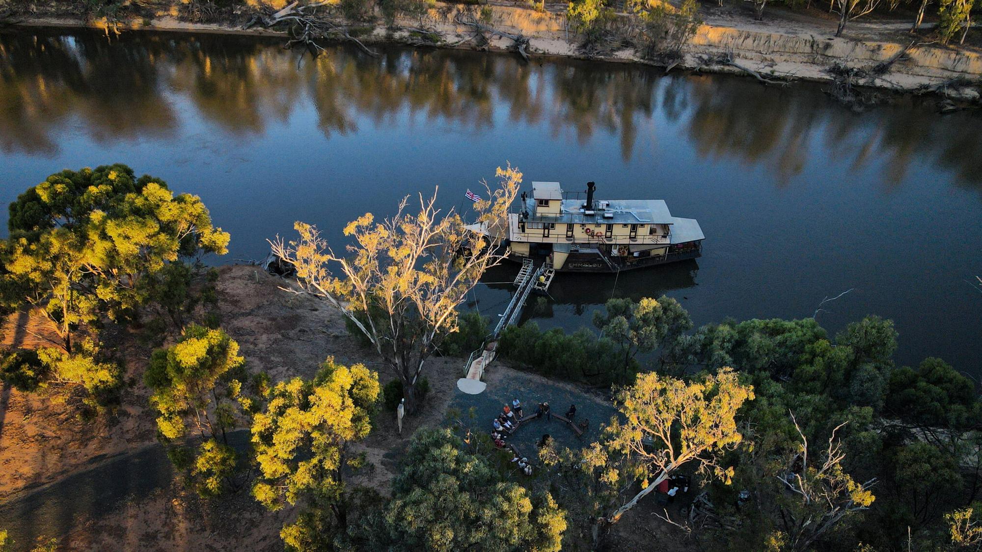 River Cruise on Murray River, Australia An aerial view of a paddlewheel riverboat moored along the Murray River in Australia, with passengers gathered on the sandy bank near the gangway. Golden-lit eucalyptus trees frame the scene, reflecting in the calm blue water, showcasing a scenic inland waterway cruise experience.