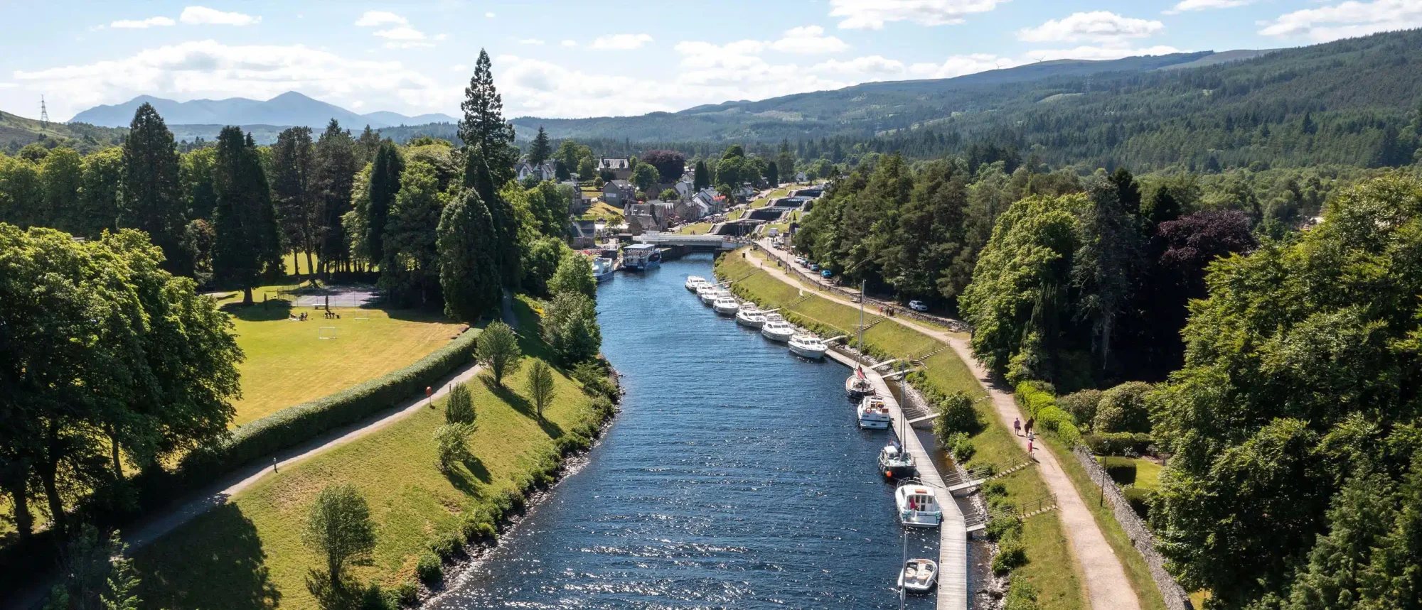 Scenic River Valley in Caledonia with Moored Boats An aerial view of a picturesque river valley lined with lush green forests, manicured lawns, and