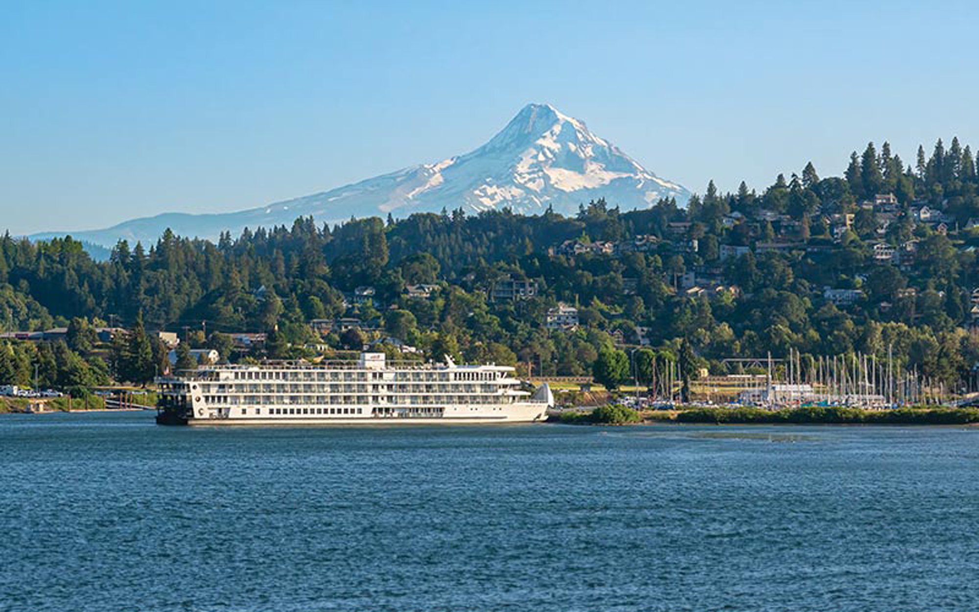 American Cruise Lines River Ship on Columbia River A multi-deck white riverboat navigates calm blue waters with snow-capped Mount Hood dominating the background, surrounded by dense evergreen forests and a hillside community nestled among the trees in the Pacific Northwest.