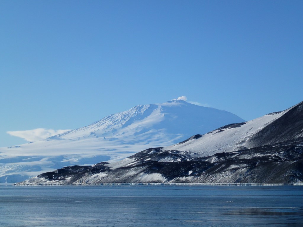 Snow-Capped Volcano Over Frozen Lake A majestic snow-covered stratovolcano rises above a partially frozen lake, with dark volcanic