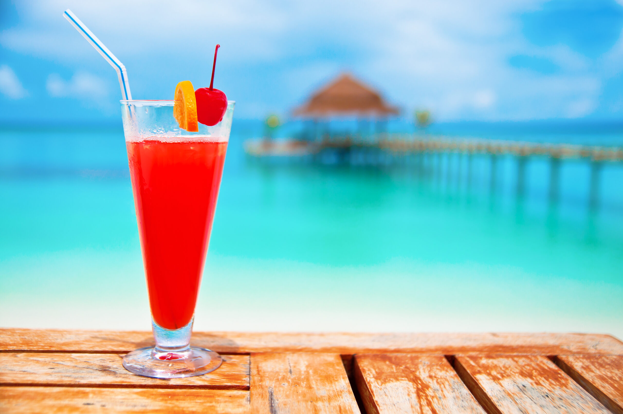 Tropical Beach Cocktail with Ocean View A vibrant red cocktail with ice, garnished with a maraschino cherry and orange slice, sits on a wooden deck overlooking turquoise tropical waters. In the soft-focus background, a thatched-roof beach bungalow and wooden pier are visible against a clear blue sky, evoking a luxurious tropical resort or cruise destination setting.
