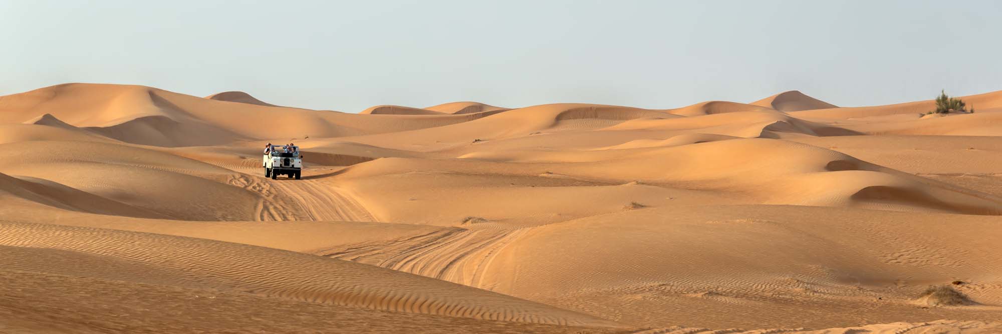 4×4 SUV Desert Safari in Dubai A vintage open-top 4x4 SUV traverses the golden sand dunes of the Dubai desert, navigating the undulating landscape with multiple sand dunes visible under clear skies. This captures the adventurous desert safari experience popular among tourists visiting the United Arab Emirates.