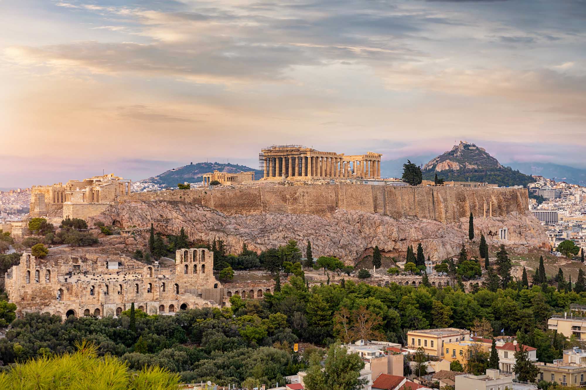 Acropolis at Sunset Over Athens A stunning panoramic view of the Acropolis in Athens, Greece, showcasing the iconic Parthenon temple complex illuminated by golden sunset light atop a rocky outcrop, with the historic Plaka district, Lycabettus Hill, and modern Athens sprawling below under a dramatic cloudy sky.
