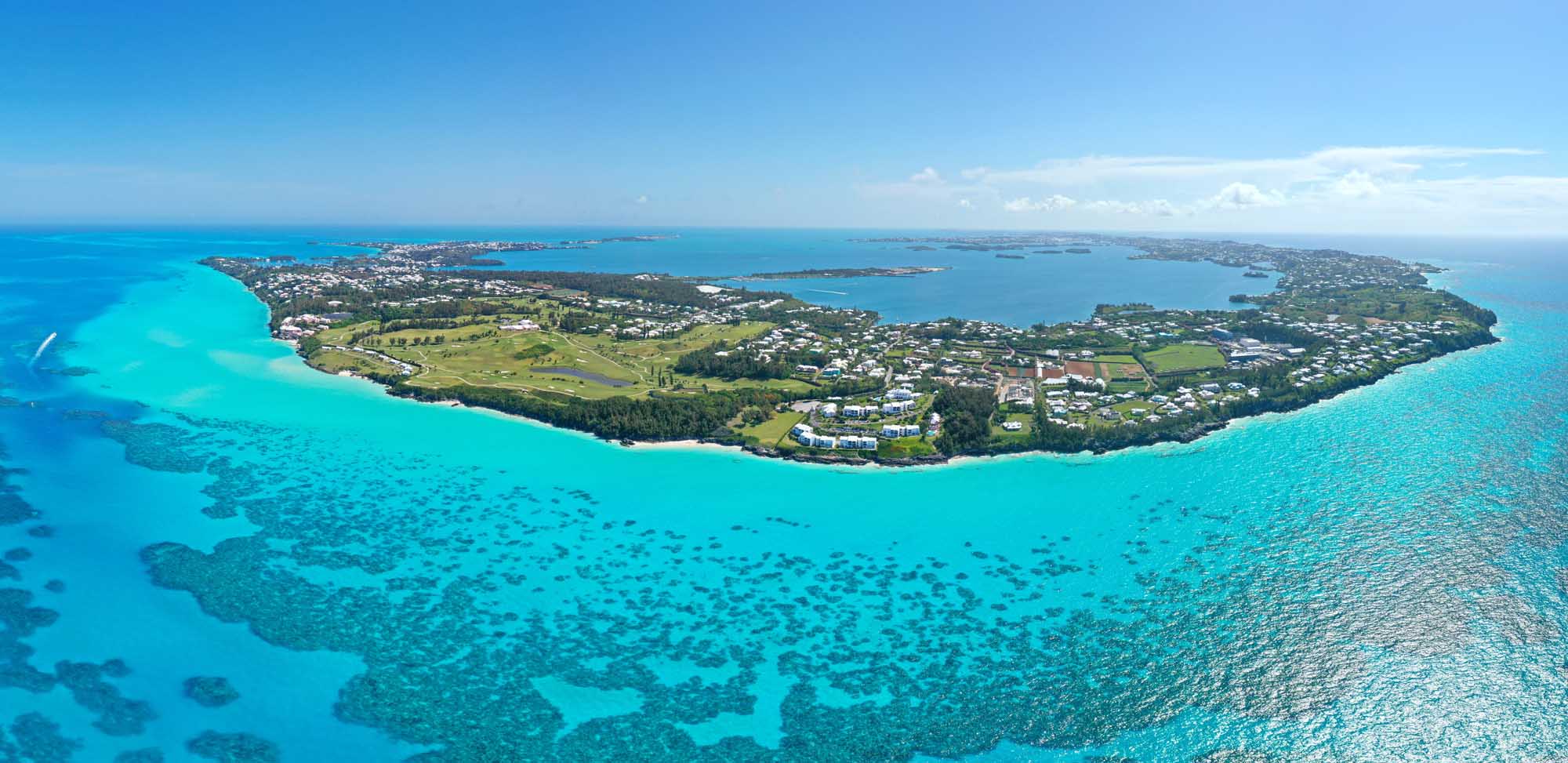 Aerial View of Bermuda Island An aerial photograph of Bermuda island surrounded by stunning turquoise Atlantic Ocean waters, showcasing the island's lush green vegetation, residential areas, and pristine beaches. The image captures the island's distinctive crescent shape with clear views of the shallow turquoise waters and darker blue deeper ocean.