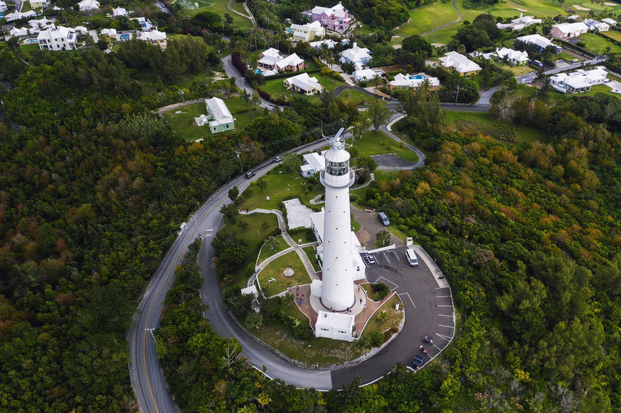 Aerial View of Bermuda Lighthouse and Coastal Community An aerial photograph of a distinctive white lighthouse with its accompanying keeper's residence situated on a promontory in Bermuda, surrounded by lush green vegetation, winding coastal roads, and residential properties. The image showcases the island's verdant landscape, curving waterways, and well-maintained suburban homes characteristic of Bermuda's affluent communities.