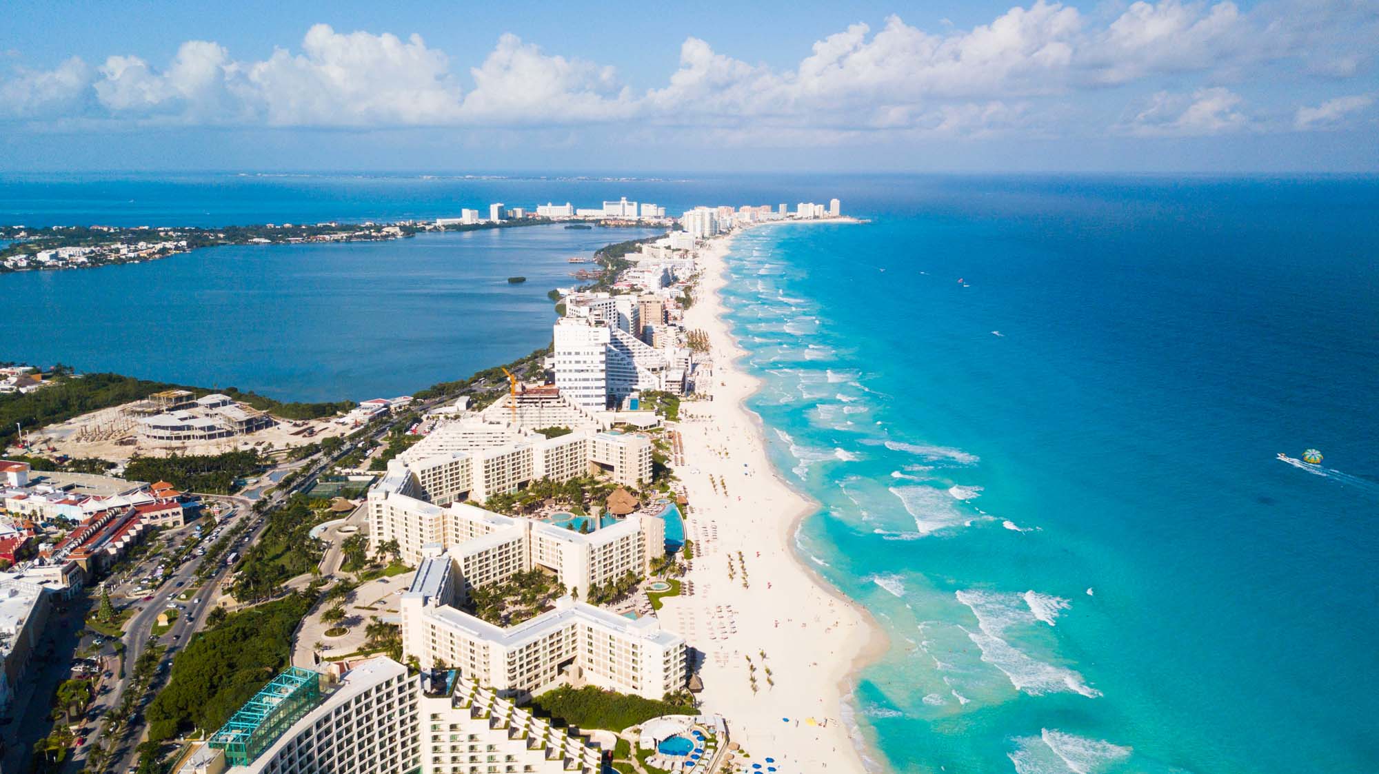 Aerial View of Cancun Beach Resort Strip An aerial photograph of Cancun's famous hotel zone showing a narrow strip of white sand beach lined with high-rise resort hotels, turquoise Caribbean waters, and the lagoon on the opposite side. The image captures the distinctive crescent shape of the developed tourist area with clear blue skies and calm waters.
