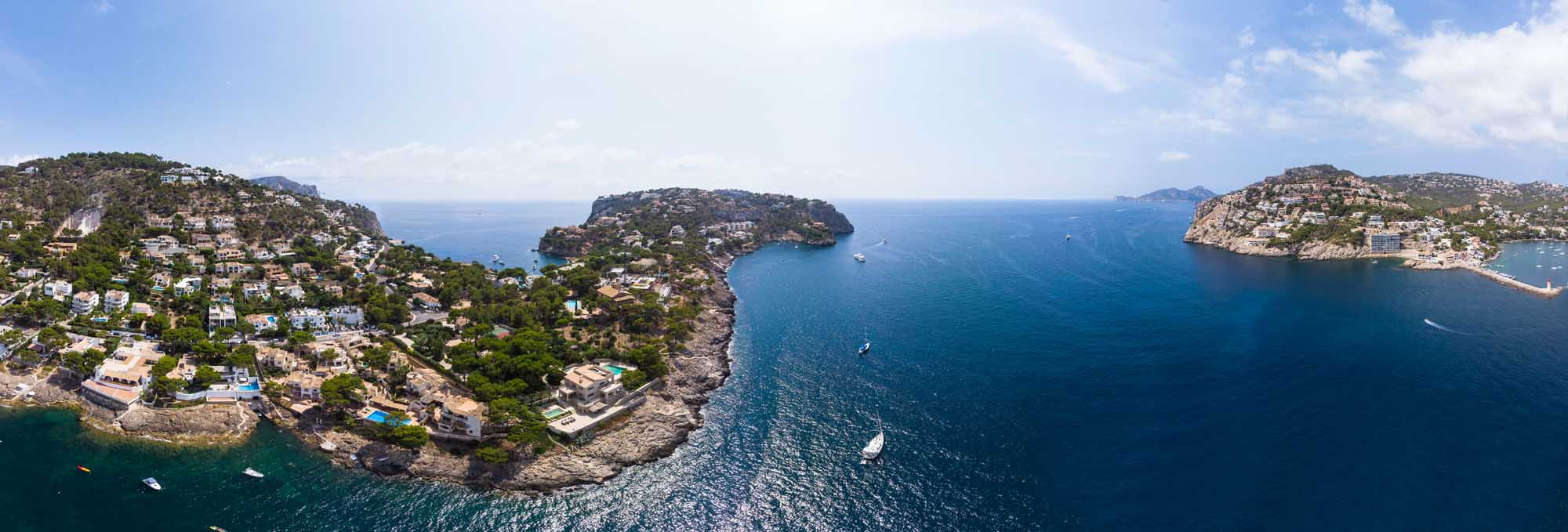 Aerial View of Port d’Andratx, Mallorca A stunning aerial photograph of Port d'Andratx in Mallorca, Balearic Islands, showcasing dramatic rocky coves, crystal-clear turquoise waters, luxury Mediterranean villas nestled on hillsides, and numerous moored yachts. The natural harbor is surrounded by lush vegetation and rocky peninsulas, with smaller neighboring islands visible in the distance.