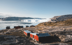 Arctic Cabin with Icebergs and Glacier A rustic orange-red cabin sits on a rocky Arctic shore overlooking a pristine fjord filled with icebergs and glaciers in the distance. The stark, barren landscape and massive ice formations exemplify the raw beauty of Arctic exploration and adventure travel.