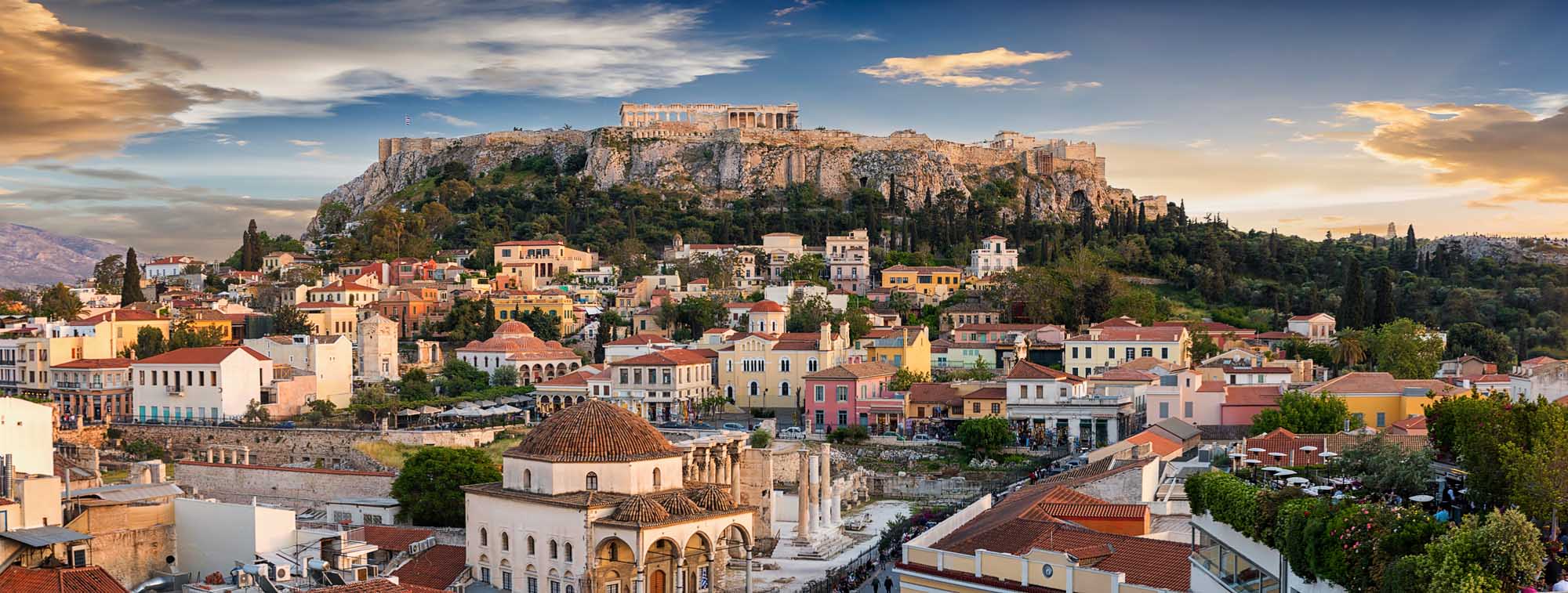 Athens Acropolis and Old Town at Sunset A panoramic view of Athens' historic old town with colorful neoclassical buildings in the foreground and the illuminated Parthenon Temple atop the Acropolis in the background, captured during golden hour sunset with dramatic clouds overhead.