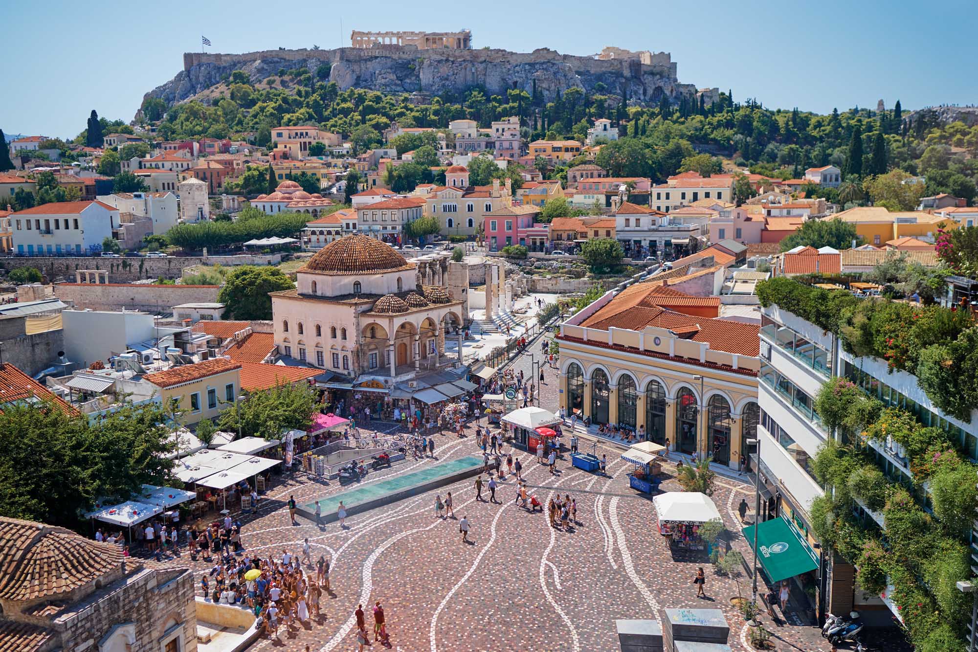 Athens Monastiraki Square with Acropolis Overlooking A vibrant daytime aerial view of Athens' historic Monastiraki Square featuring the distinctive domed mosque, colorful neoclassical buildings, bustling marketplace, and the iconic Acropolis perched dramatically on the hilltop in the background under clear blue skies.