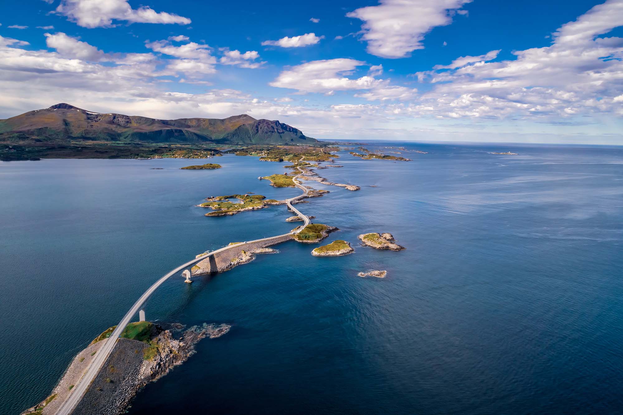 Atlantic Ocean Road, Norway Aerial View An aerial photograph of the Atlantic Ocean Road (Atlanterhavsveien) in Norway, showcasing a dramatic serpentine causeway connecting small islands across the blue Atlantic waters. The road winds through a stunning coastal landscape with mountains visible in the background and pristine blue skies dotted with white clouds.