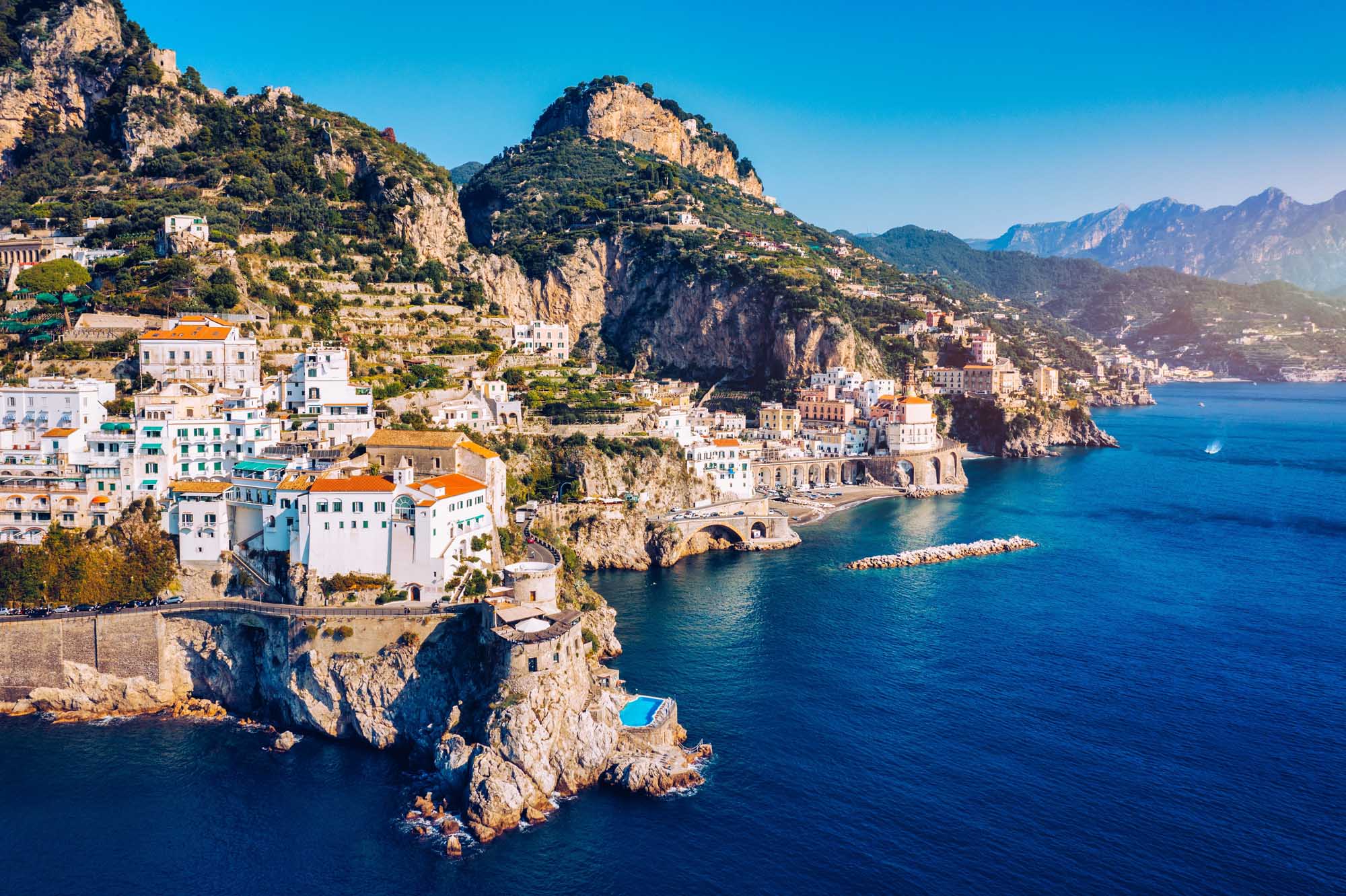 Atrani Village on the Amalfi Coast An aerial view of Atrani, a picturesque coastal village clinging to dramatic cliffsides along Italy's Amalfi Coast. Colorful pastel-hued buildings cascade down the rocky terrain toward crystal-clear turquoise Mediterranean waters, with the surrounding mountains and neighboring villages visible in the distance.