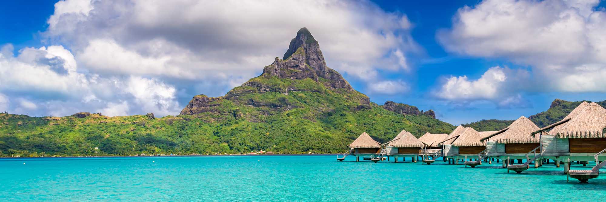 Bora Bora Island with Overwater Bungalows A stunning tropical paradise view of Bora Bora Island in French Polynesia, featuring iconic thatched-roof overwater bungalows in turquoise lagoon waters, with the dramatic volcanic Mount Otemanu peak rising majestically in the background under a bright blue sky with white clouds.