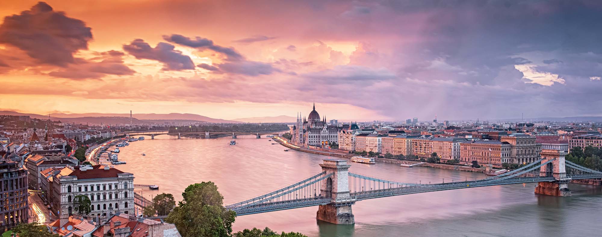 Budapest Sunset Over the Danube River A stunning panoramic view of Budapest at golden hour, featuring the iconic Chain Bridge spanning the Danube River with the Hungarian Parliament Building illuminated on the right bank. The cityscape is bathed in warm orange and pink hues as the sun sets behind dramatic clouds, reflecting beautifully across the water.