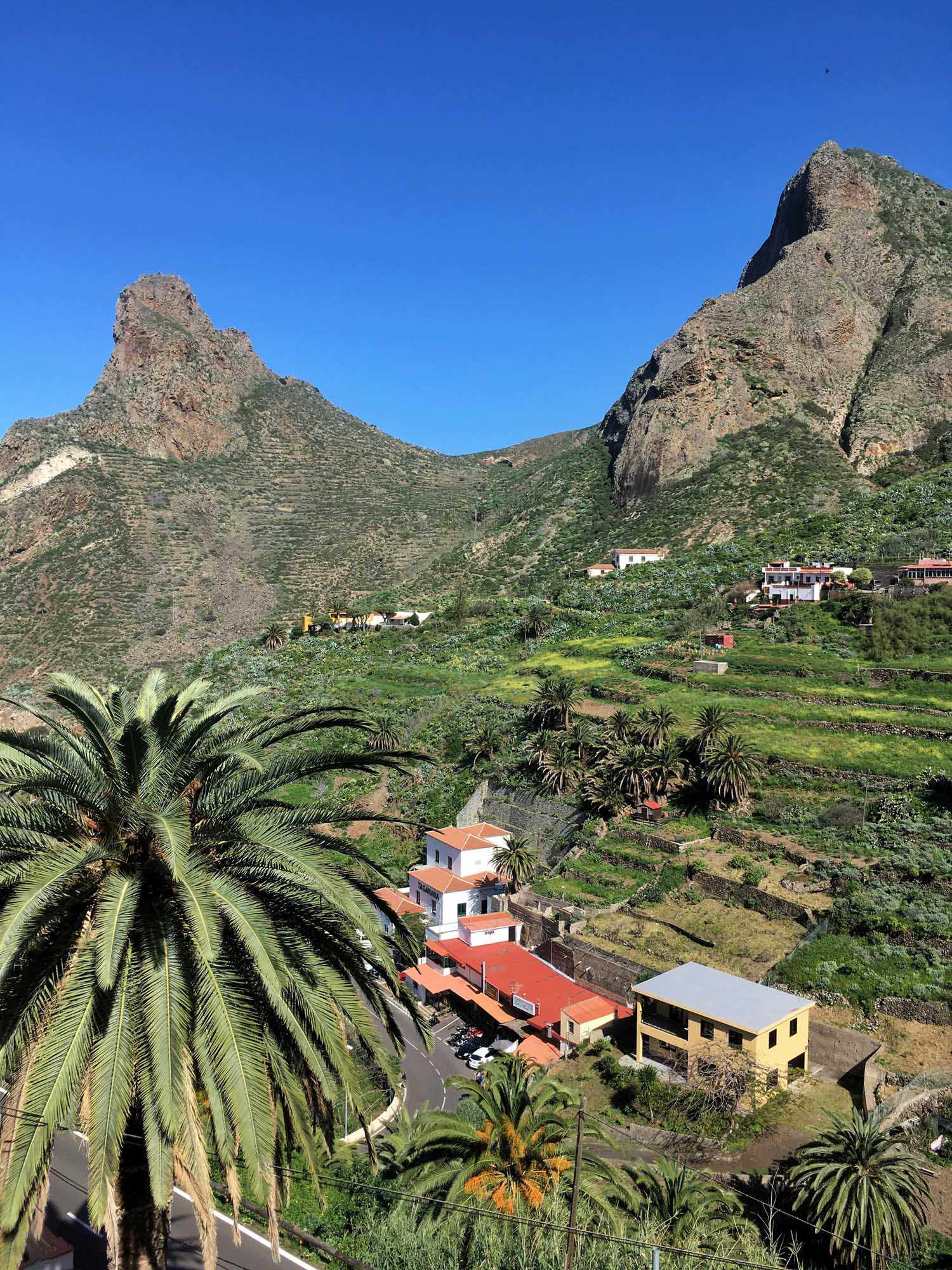 Canary Islands Mountain Village Valley A picturesque valley settlement nestled between dramatic volcanic peaks in the Canary Islands, featuring traditional white and colorful buildings with terracotta roofs, lush green agricultural terraces, palm trees, and a clear blue sky. The landscape showcases the unique combination of arid mountainous terrain and verdant valley cultivation typical of the archipelago's diverse geography.