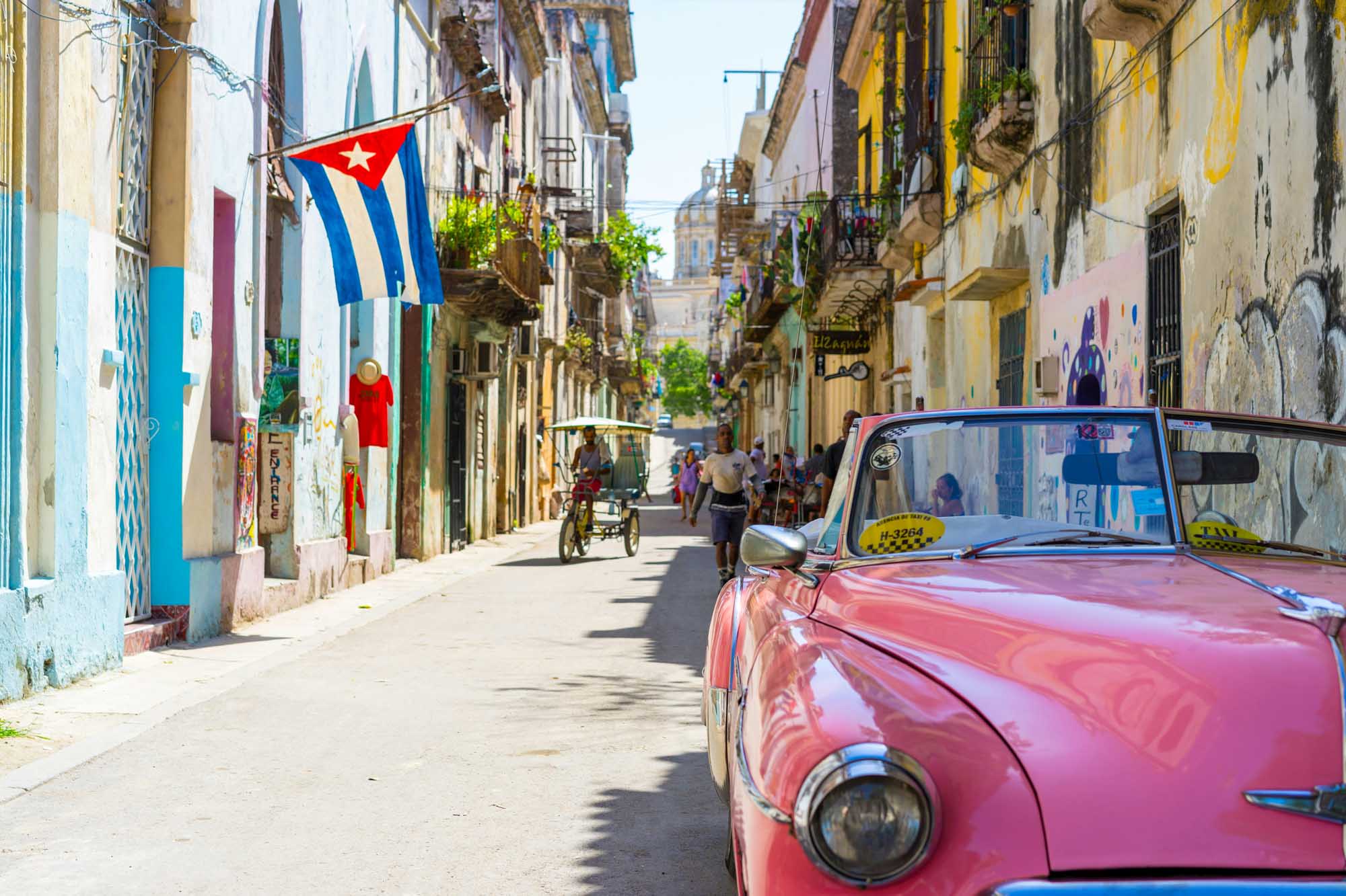 Classic Car and Cuban Flag in Havana Street A vibrant street scene in Havana, Cuba featuring a pink vintage American car parked along a colorful colonial building, with a Cuban flag hanging overhead. The image captures the iconic charm of Old Havana with its weathered architecture, narrow cobblestone streets, traditional horse-drawn cart, and the distinctive aesthetic that makes Cuba a unique Caribbean destination.