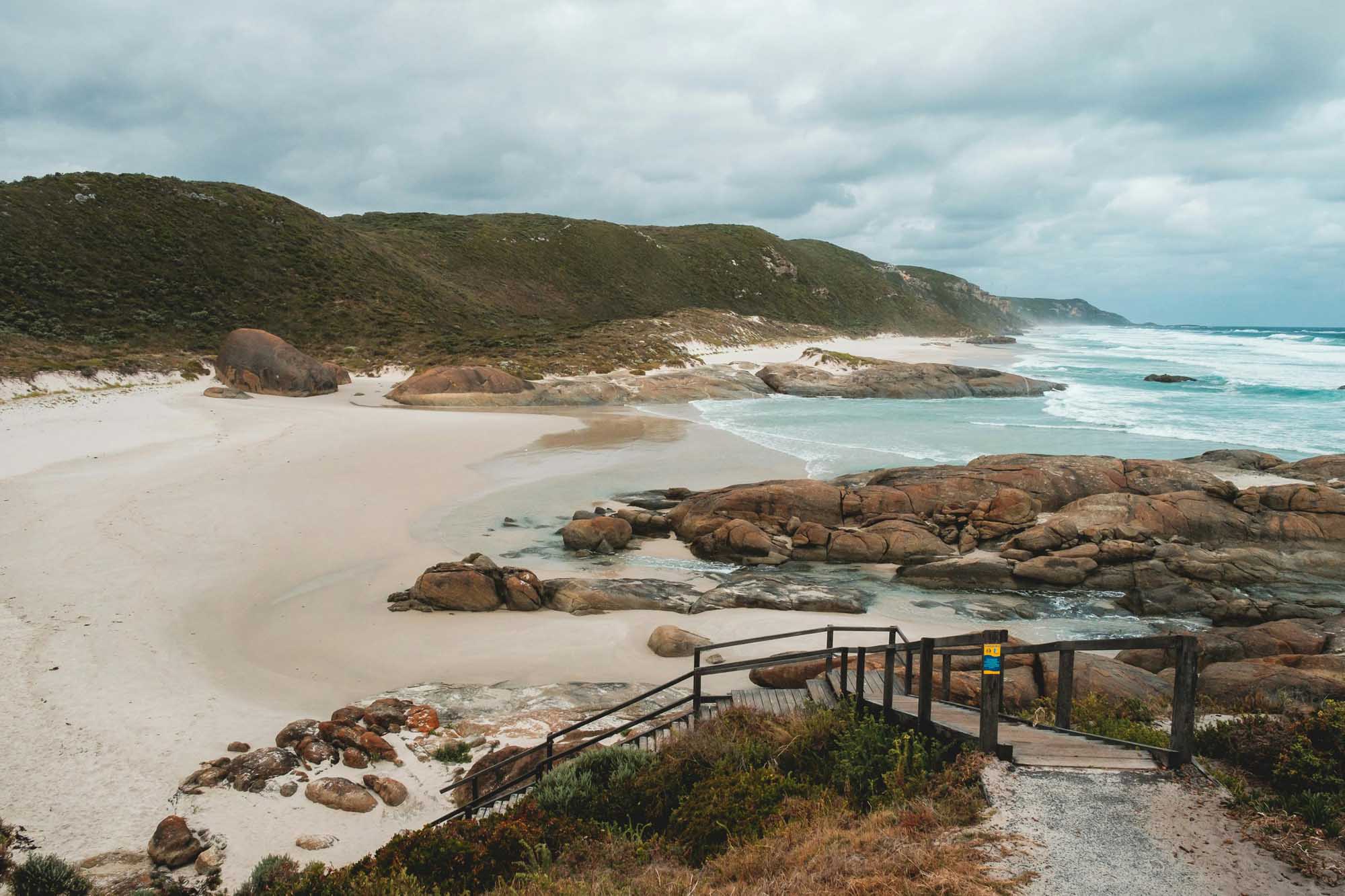 Coastal Beach with Rocky Outcrops and Wooden Boardwalk A picturesque coastal landscape featuring a pristine white sand beach framed by distinctive granite rock formations, accessible via a wooden boardwalk. Turquoise waters, lush green hillsides, and dramatic cloud-filled skies complete this scenic seaside destination.