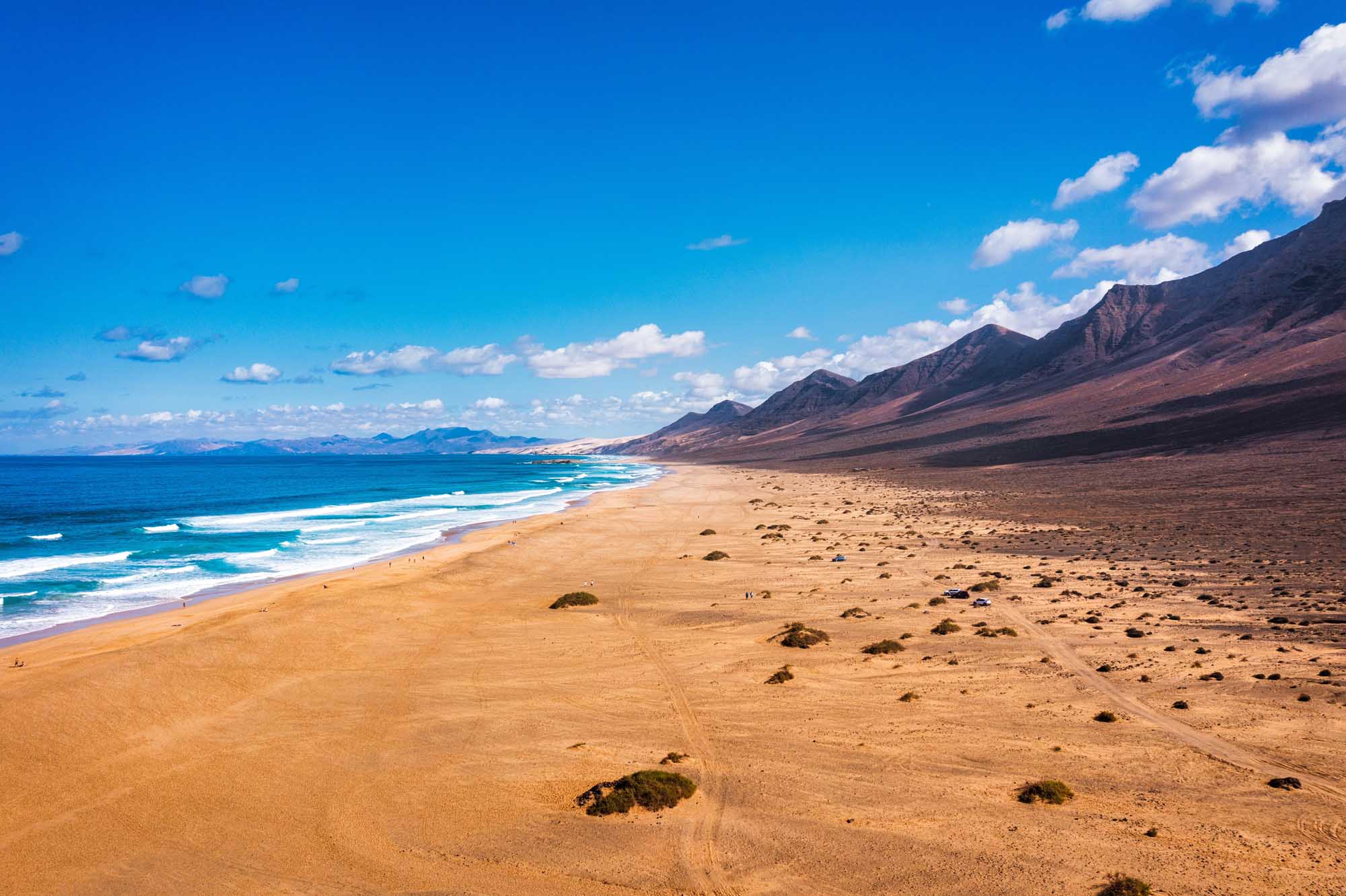 Cofete Beach with Volcanic Hills, Fuerteventura A stunning panoramic view of Cofete beach in Fuerteventura, Canary Islands, featuring golden sand stretching along the Atlantic Ocean with dramatic volcanic mountains rising in the background under a brilliant blue sky with scattered clouds.