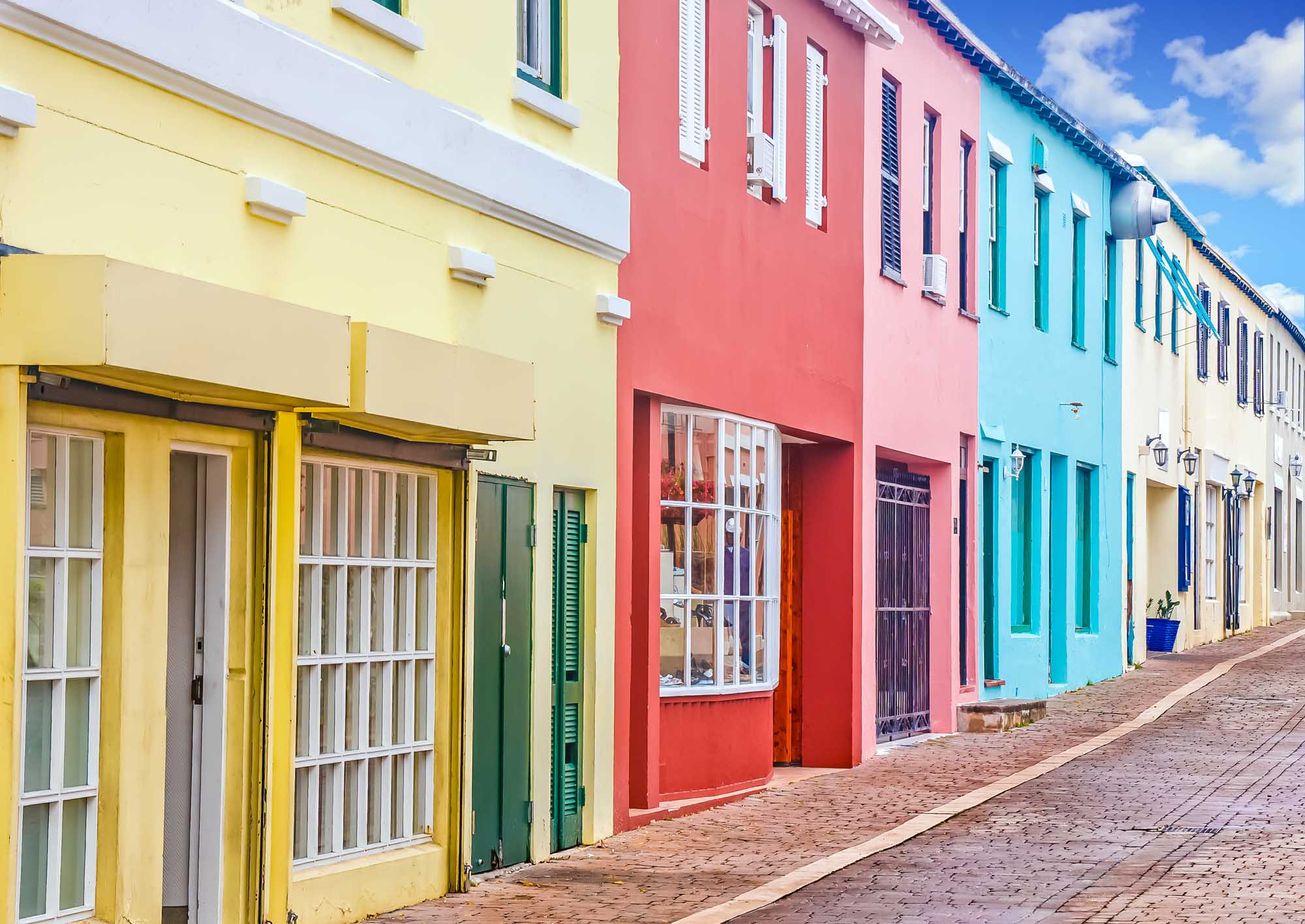 Colorful Colonial Architecture in Bermuda A picturesque street lined with vibrant pastel-colored colonial buildings featuring yellow, pink, turquoise, and cream facades. White shuttered windows and traditional wooden doors characterize the historic architecture, with a brick-paved street leading through this charming residential or commercial district typical of Bermuda's distinctive aesthetic.