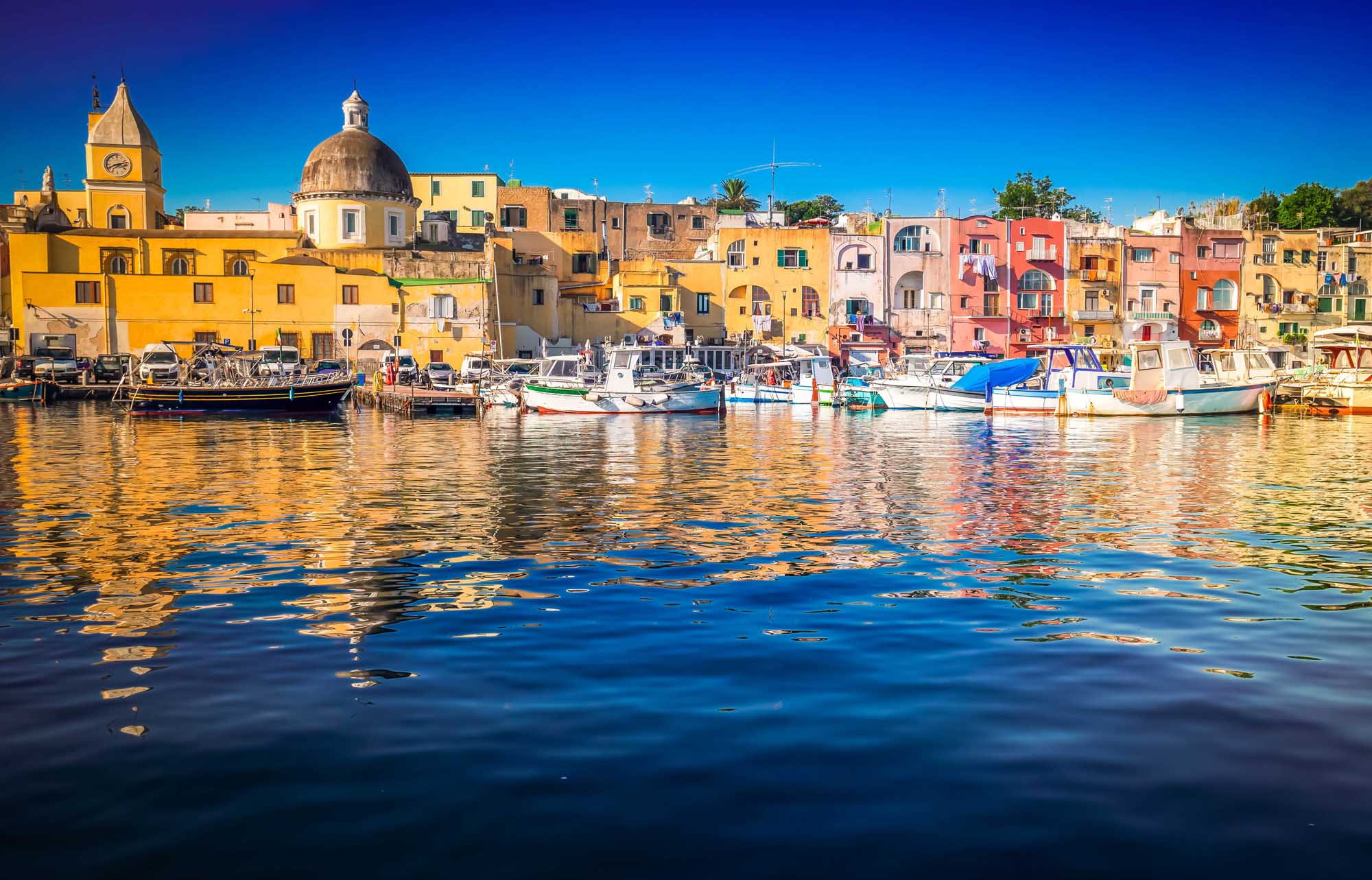 Colorful Marina of Procida Island, Italy A vibrant Mediterranean harbor scene featuring the iconic Marina Grande of Procida Island, with densely packed pastel-colored buildings in yellow, pink, orange, and cream tones reflecting in the calm blue waters. Traditional fishing boats are moored along the waterfront, with a prominent domed church visible among the historic architecture.