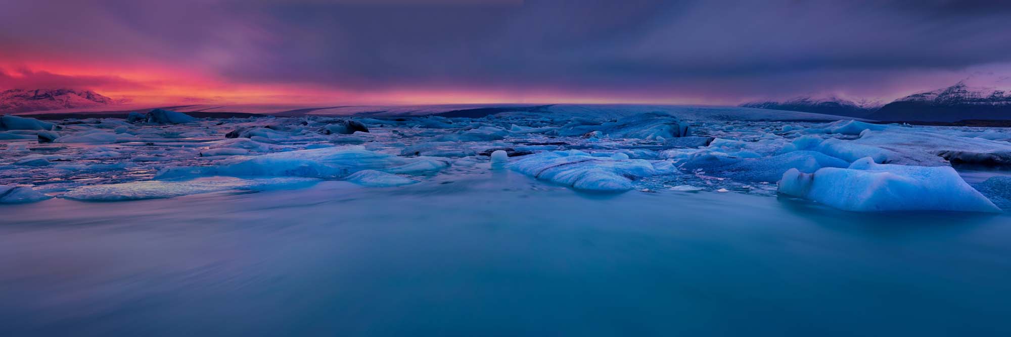 Crimson Afterglow at Jokulsarlon Glacier Lagoon A stunning twilight panorama of Iceland's Jokulsarlon glacier lagoon, featuring icebergs floating in crystalline turquoise water beneath a dramatic sunset sky painted in shades of crimson, purple, and deep blue. The surreal landscape captures the raw beauty of Iceland's glacial region during the magical golden hour.