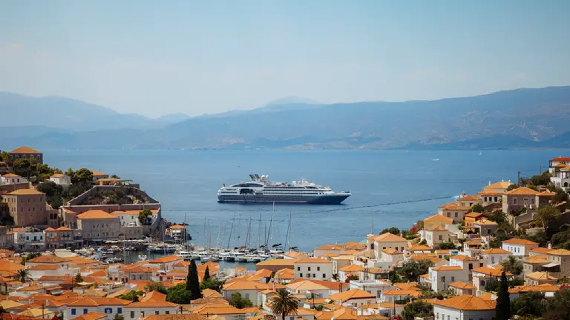 Cruise Ship Anchored at Hydra, Greece A large modern cruise ship anchored in the crystalline Mediterranean waters off the picturesque Greek island of Hydra, with traditional whitewashed buildings featuring orange-tiled roofs cascading down the hillside, sailboats moored in the harbor, and the mainland mountains visible across the water in the distance.