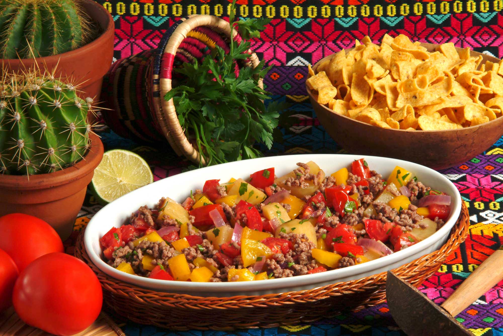 Cuban Picadillo with Traditional Styling A vibrant presentation of Cuban picadillo habanero served in a white ceramic bowl, featuring seasoned ground meat with diced potatoes, red and yellow peppers, and fresh cilantro garnish. The dish is artfully arranged on a colorful woven placemat alongside complementary items including fresh lime, a decorative ceramic pitcher, potted cactus, crispy tortilla chips, and fresh tomatoes, creating an authentic and appetizing Mexican-Cuban culinary scene.