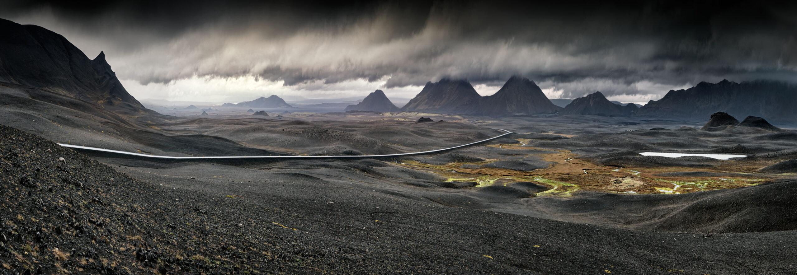 Dramatic Icelandic Highland Landscape with Mountains A sweeping panoramic view of Iceland's stark volcanic highlands, featuring a winding glacial river cutting through dark lava fields, dramatic jagged mountain peaks in the distance, and moody storm clouds overhead. The landscape showcases the raw, otherworldly terrain characteristic of Iceland's interior, with patches of golden vegetation dotting the desolate terrain.