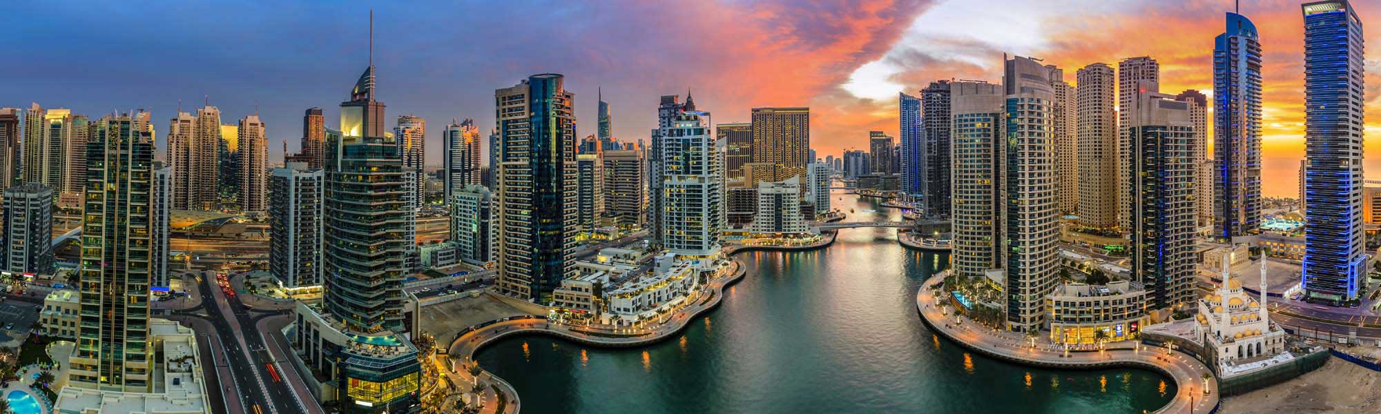 Dubai Marina Skyline at Sunset A stunning panoramic view of Dubai Marina at golden hour, showcasing the iconic waterfront development with modern high-rise residential and commercial towers reflected in the calm marina waters. The vibrant sunset sky in shades of orange and gold illuminates the contemporary architecture and bustling waterfront promenade lined with restaurants and entertainment venues.