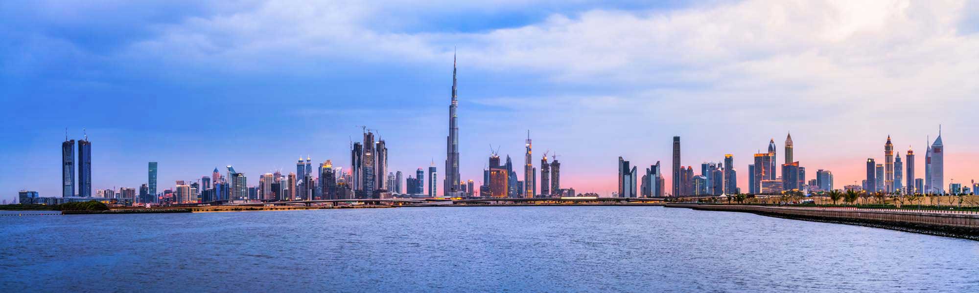 Dubai Skyline at Sunset Over the Creek A panoramic view of Dubai's iconic skyline featuring the Burj Khalifa and modern skyscrapers reflected in the calm waters of Dubai Creek during golden hour, with palm trees lining the waterfront.