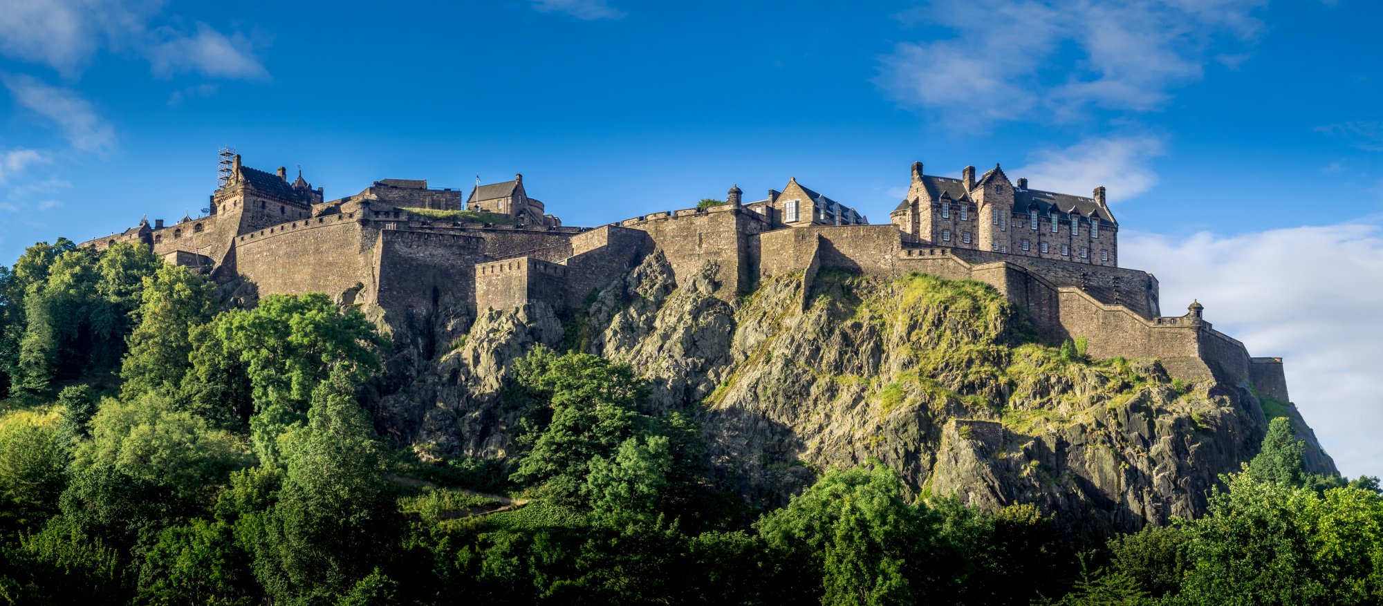Edinburgh Castle Perched on Volcanic Rock Edinburgh Castle stands majestically atop a dramatic volcanic crag overlooking the city of Edinburgh, Scotland. The historic fortress features distinctive stone architecture with turrets and defensive walls, surrounded by lush green vegetation on the steep rocky outcrop.