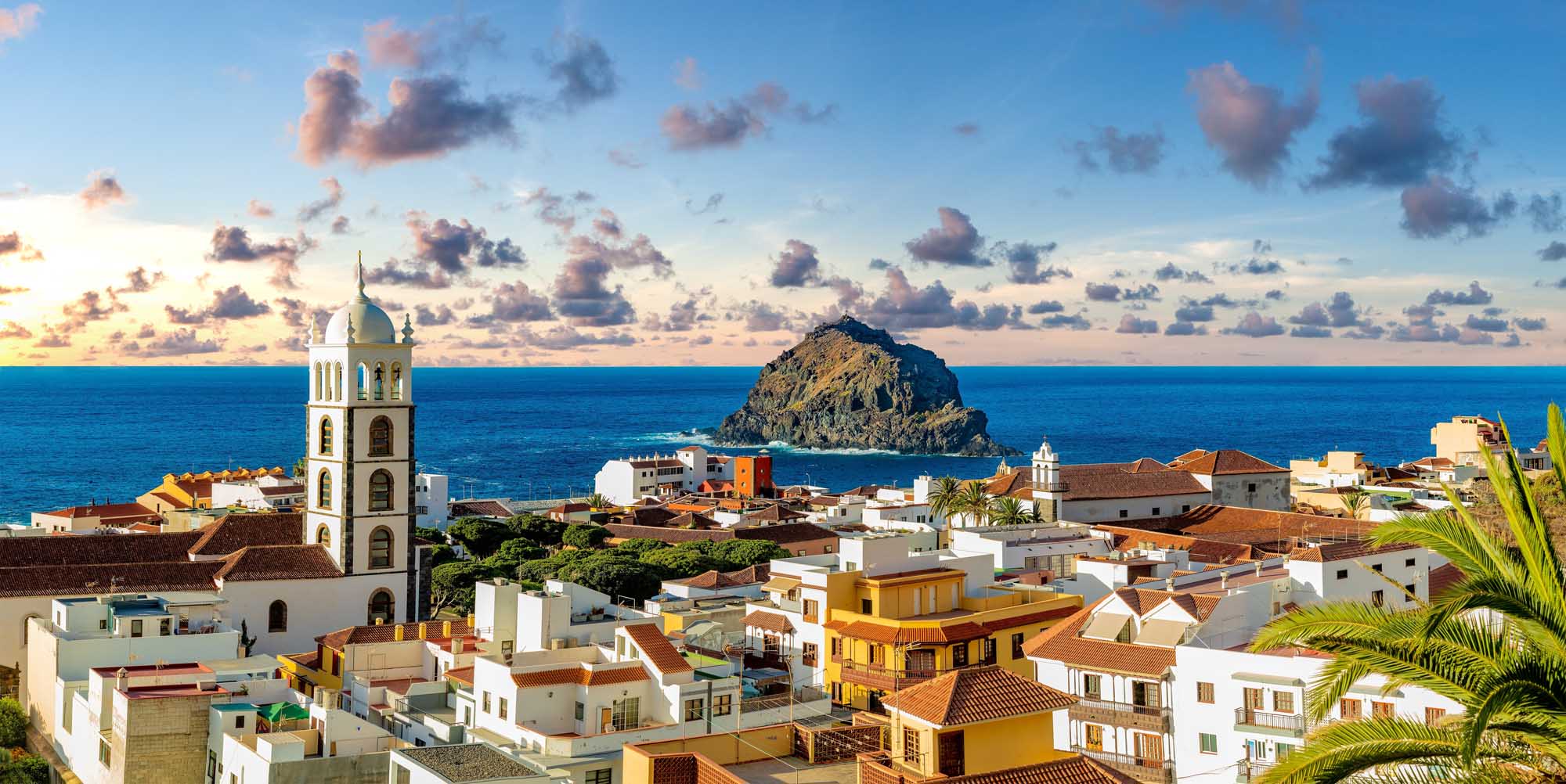 Garachico Village Coastal Panorama, Tenerife A picturesque panoramic view of the historic coastal village of Garachico in Tenerife, Canary Islands, featuring colorful whitewashed and yellow buildings with terra cotta roofs, a prominent church bell tower, and a distinctive dark volcanic rock formation rising from the turquoise Atlantic Ocean under a dramatic cloud-filled sky at sunset.