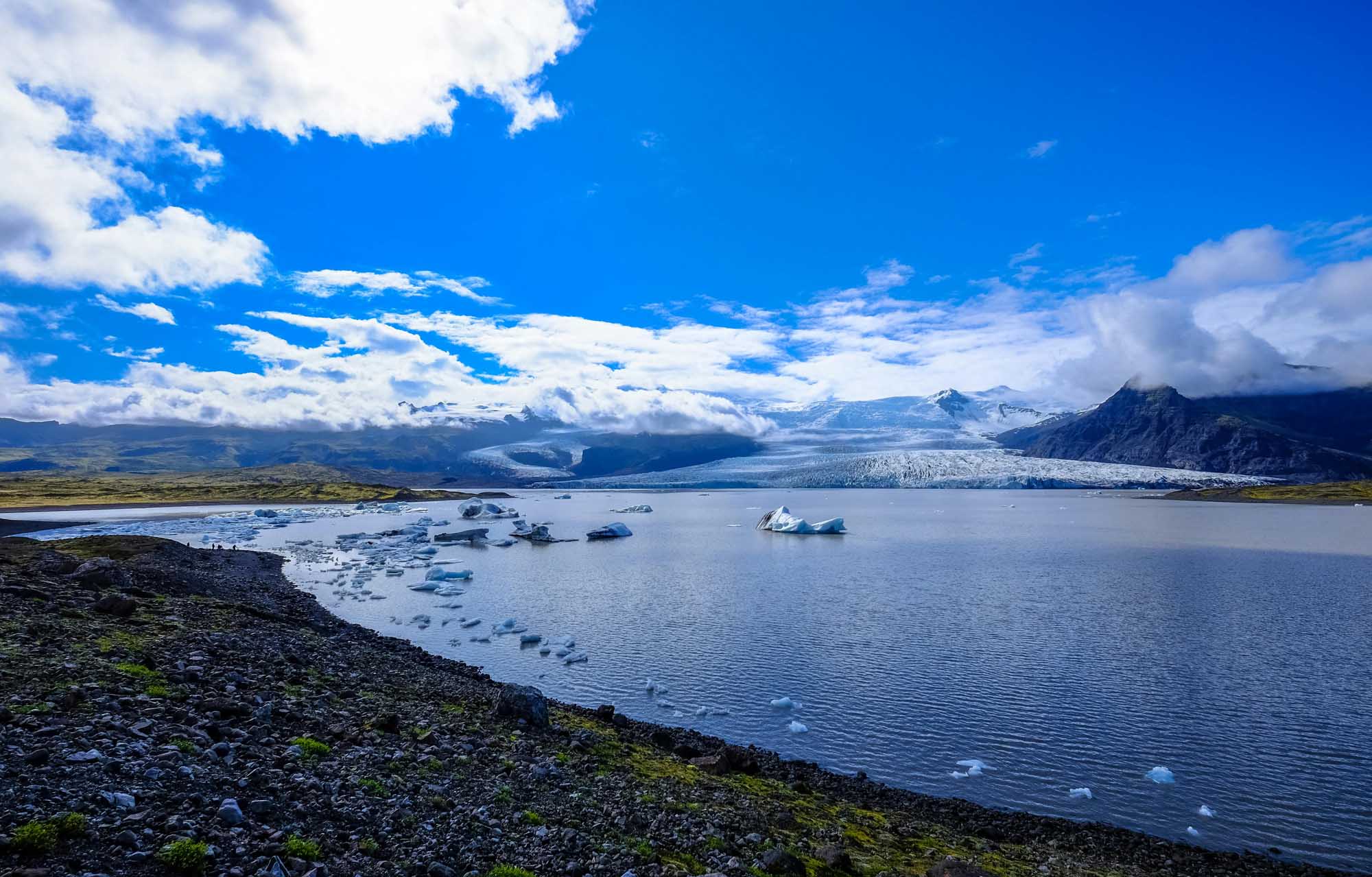 Glacial Lake with Icebergs in Iceland A stunning glacial landscape featuring a pristine blue lake dotted with floating icebergs, with a massive glacier visible in the distance against snow-capped mountains and a bright blue sky. The foreground shows dark volcanic rock shoreline with patches of moss, typical of Iceland's dramatic natural scenery.